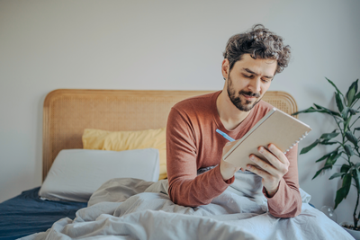 Mann sitzt im Bett und schreibt etwas in ein Notizbuch