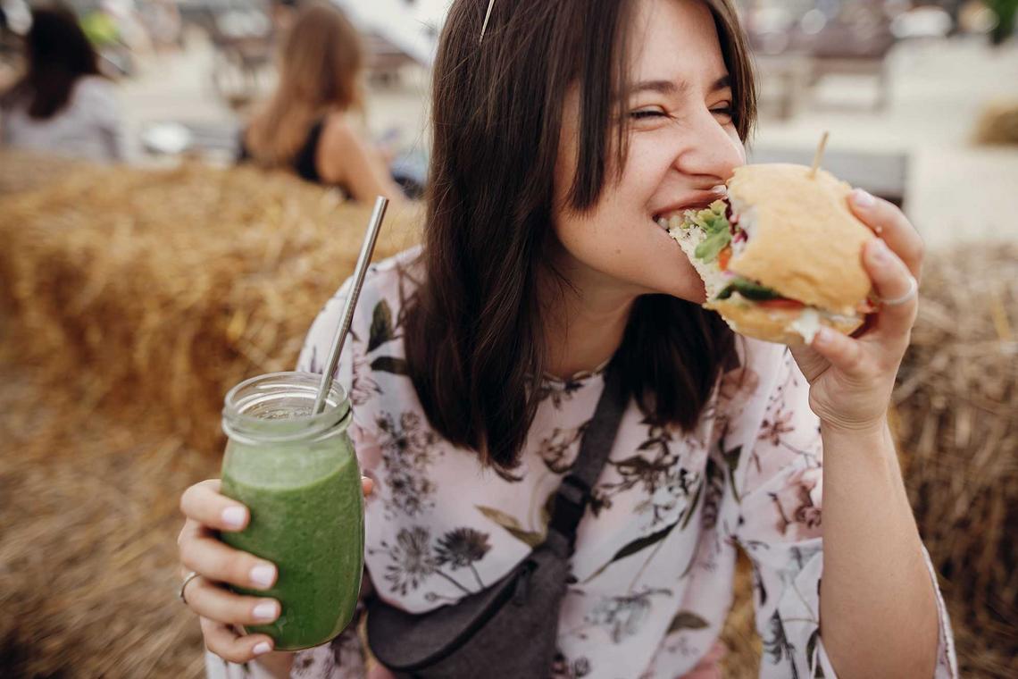Bildnachweis: © stock.adobe.com / sonyachny Junge Frau hält einen Smoothie in der Hand und beißt in einen veganen Burger